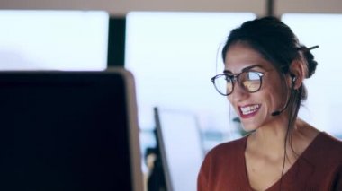 Ofiste oturmuş kulaklıkla konuşurken bilgisayarla çalışan genç ve güzel bir iş kadınının fotoğrafı..