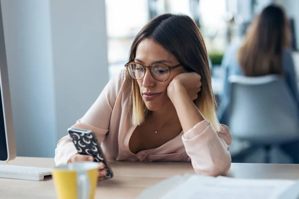 Shot of boring young business woman sending messages with mobile phone while sitting in modern startup office.