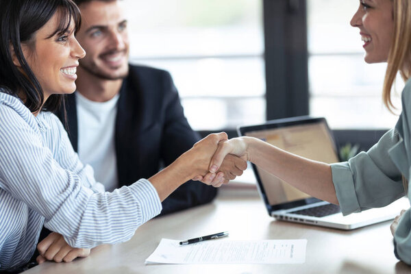 Снимок привлекательного агента по недвижимости, пожимающего руку молодой паре после подписания контракта в офисе.