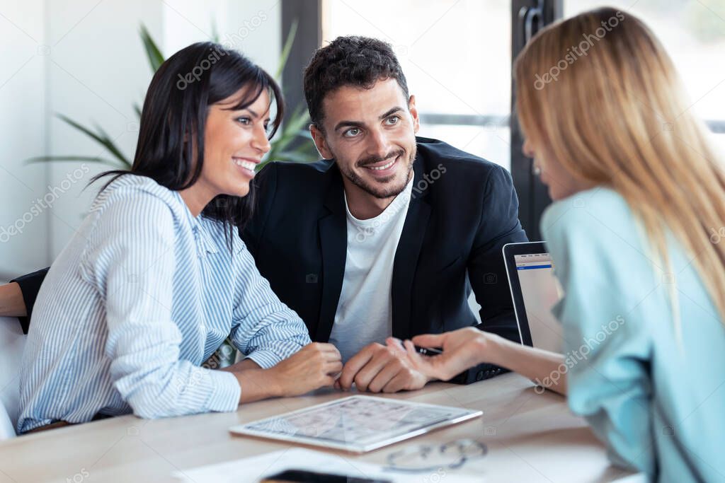 Shot of beautiful real-estate agent showing house plans on electronic tablet while talking to the couple about buying the house in the office.