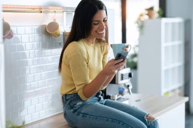 Evdeki mutfakta oturmuş kahve içerken akıllı telefon kullanan çekici bir genç kadın..