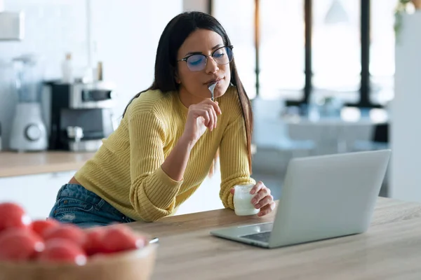 自宅でキッチンに座ってヨーグルトを食べている間 自信を持って若い女性のコンピュータで作業のショット — ストック写真