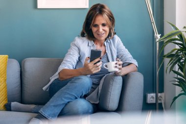 Evdeki kanepede oturup kahve içerken oldukça olgun bir kadının cep telefonunu kullanması..