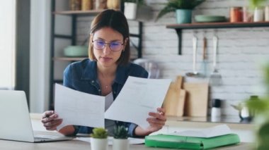 Evdeki mutfakta faturalara ve belgelere bakarken bilgisayarla çalışan güzel bir iş kadınının videosu..