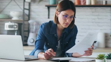 Evdeki mutfakta faturalara ve belgelere bakarken bilgisayarla çalışan güzel bir iş kadınının videosu..