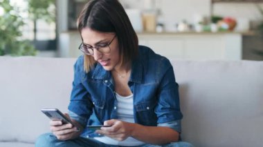 Evdeki oturma odasında cep telefonuyla online alışveriş yapmak için beyaz kredi kartı tutan güzel bir kadının videosu..