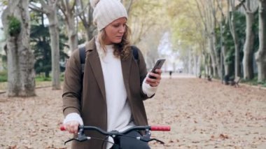Sonbaharda parkta bisiklet sürerken akıllı telefonuyla mesaj gönderen güzel bir genç kadının videosu..