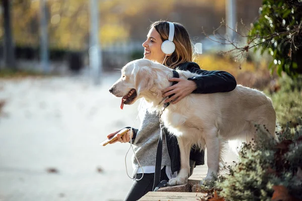 她和可爱的金发碧眼的猎犬坐在公园里 一边用智能手机听音乐 一边笑逐颜开 — 图库照片