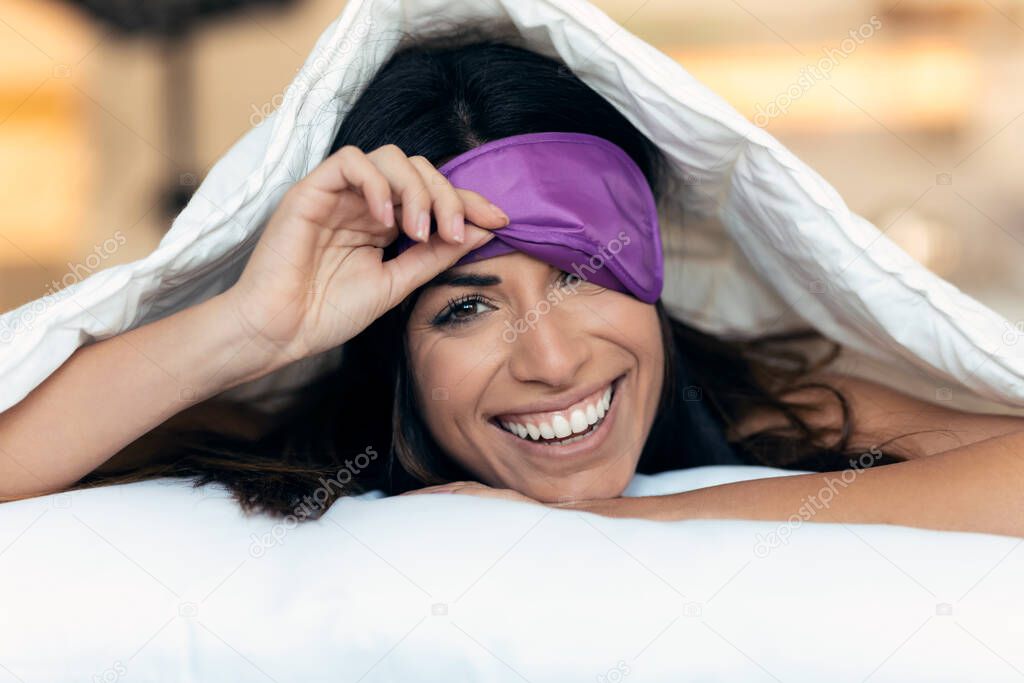 Portrait of pretty young woman pulling up sleeping mask while looking at camera after wake up in the bedroom at home.