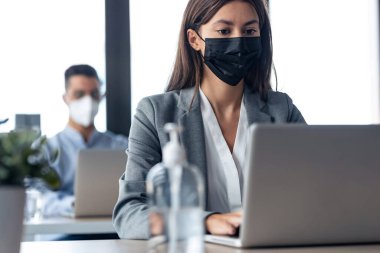 İş yerinde dizüstü bilgisayarlarla çalışırken iki çekici iş ortağının hijyenik yüz maskesi takması. Sosyal uzaklık kavramı.