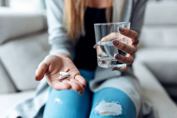 Close up of young woman taking pills while holding glass of water sitting on the couch at home.