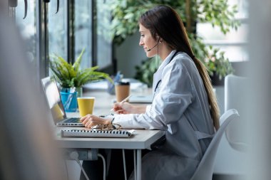 Genç bayan doktorun laptopla video görüşmesi yaparken kulaklıkla konuşurken görüntüsü..