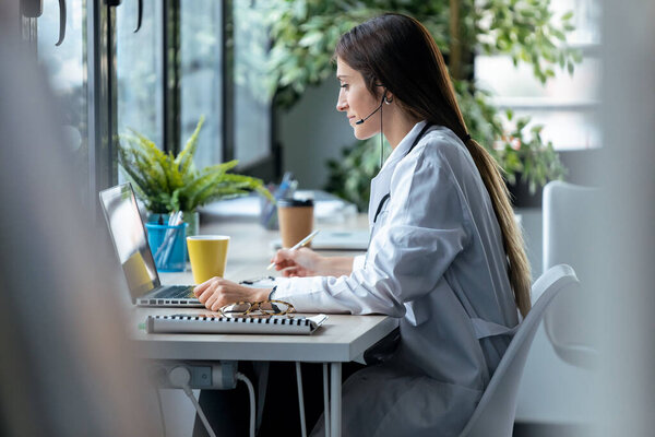 Снимок молодой женщины-врача, делающей видеозвонок с ноутбука во время разговора с наушником сидя в консультации.