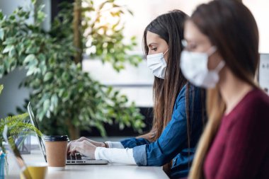İki genç iş kadınının, birlikte çalıştığı yerde dizüstü bilgisayarlarla çalışırken hijyenik yüz maskesi taktıkları bir fotoğraf.. 