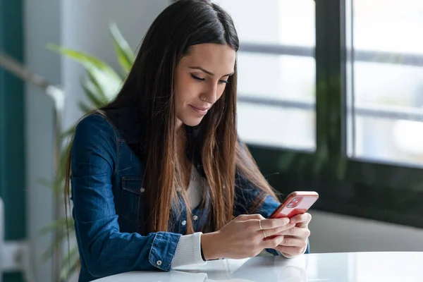 自宅のデスクで働いている間に携帯電話でメッセージを送る笑顔の若いビジネス女性のショット — ストック写真