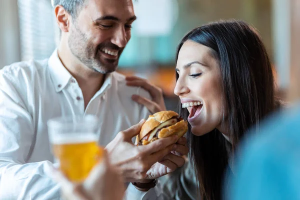 Shot of attractive man giving hamburger to his beautiful friend in the mouth while eating with their friends in the restaurant.