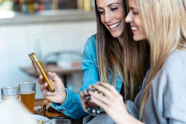 Tiro Duas Belas Jovens Amigas Comendo Hambúrgueres Enquanto Procura Telefone — Fotografia de Stock