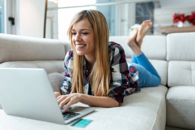 Evdeki kanepede yatarken kredi kartıyla bir şeyler ödeyen gülümseyen genç bir kadın..