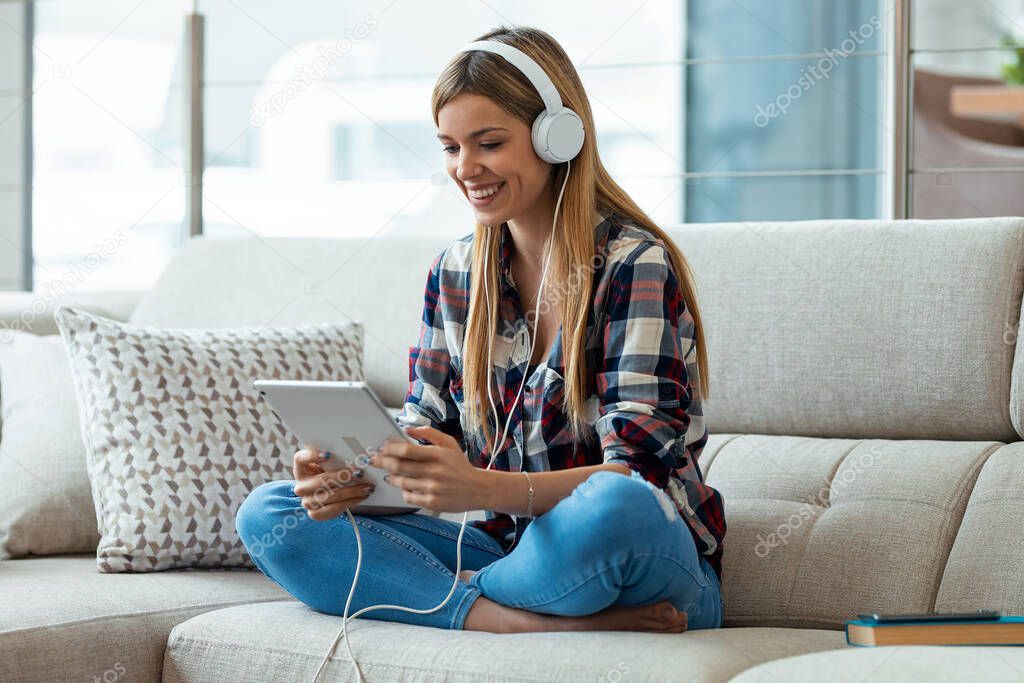 Shot of beautiful young woman listening to music with headphones while using her digital tablet sitting on couch at home.