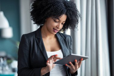Evdeki ofiste dikilirken dijital tabletini kullanan akıllı afro genç girişimci kadının portresi.