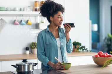 Evde yemek çubuklarıyla erişte yerken cep telefonuyla ses gönderen gülümseyen genç afro kadın..