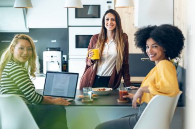 Üç zeki girişimci kadının, iş yerindeki mutfaktaki kameraya bakarken dinlenirken ve kahvaltı yaparken konuştuğu bir fotoğraf..