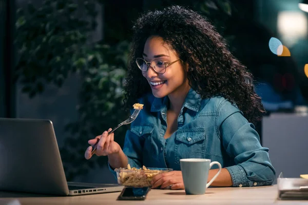 Shot of beautiful afro young business woman working with computer while eating pasta sitting in the office.