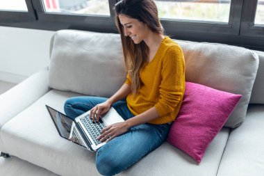 Evdeki kanepede otururken dizüstü bilgisayarıyla çalışan güzel bir genç kadının fotoğrafı..