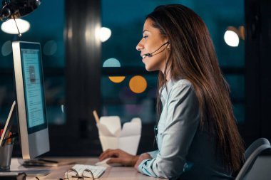 Geceleri ofiste kulaklıkla konuşurken bilgisayarla çalışan güzel bir iş kadınının görüntüsü..