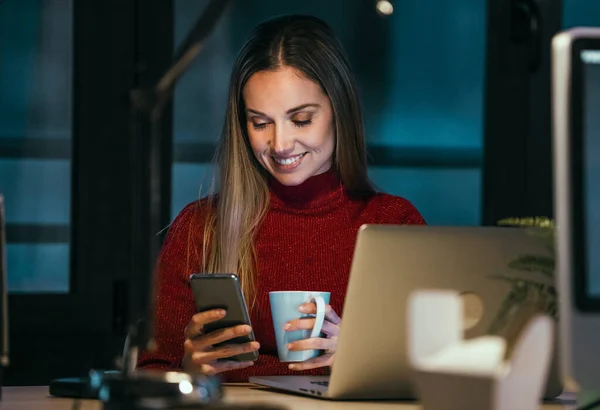 美しいビジネスの若い女性の肖像画彼女の携帯電話を使用しながら 夜にオフィスでノートパソコンで作業 — ストック写真