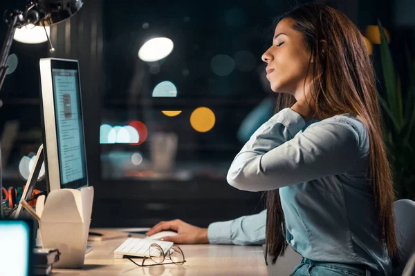 Shot of tired business woman with neck pain looking uncomfortable while working with computer in the office at night.