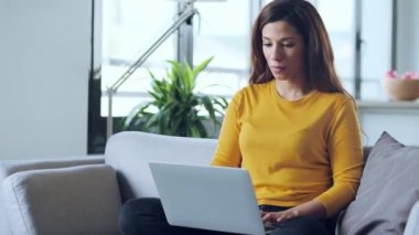 Evdeki kanepede otururken dizüstü bilgisayarıyla çalışan güzel bir genç kadının videosu..