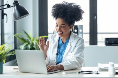 Shot of attractive afro female doctor talking while explaining medical treatment to patient through a video call with laptop in the consultation. clipart