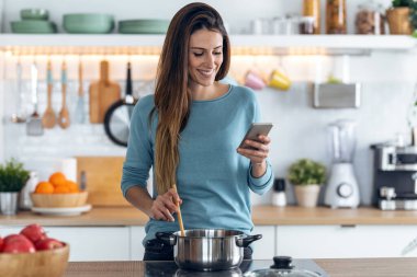 Mutfakta cep telefonunu kullanırken gülümseyen genç bir kadının sağlıklı yemek pişirmesi..