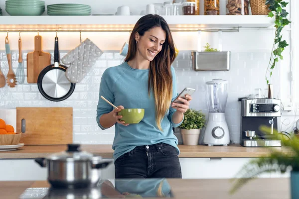 自宅の台所で箸で麺を食べながら スマートフォンでメッセージを送る美しい女性のショット — ストック写真