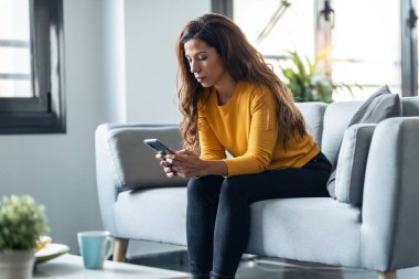 Shot of worried exhausted woman looking her smartphone while waiting for news sitting on couch at home.