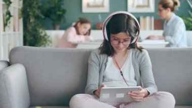 Annesi ve kız kardeşi oturma odasında çalışırken dijital tabletle müzik dinleyen küçük kızın videosu..