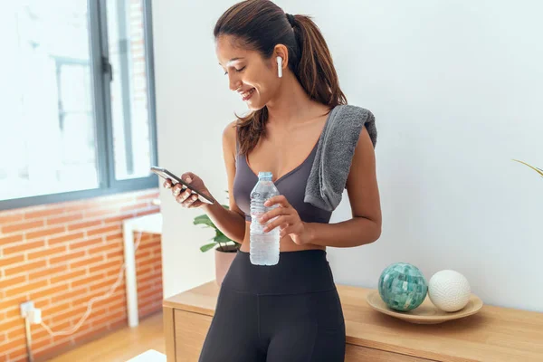 Shot of sporty young woman sending messages with her smart phone while take a break of exercise standing in living room at home.