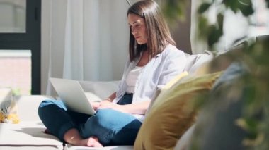 Evdeki kanepede otururken dizüstü bilgisayarıyla çalışan güzel bir genç kadının videosu..