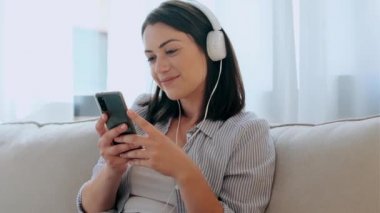 Evde kanepede otururken akıllı telefonuyla müzik dinleyen gülümseyen genç bir kadının videosu..