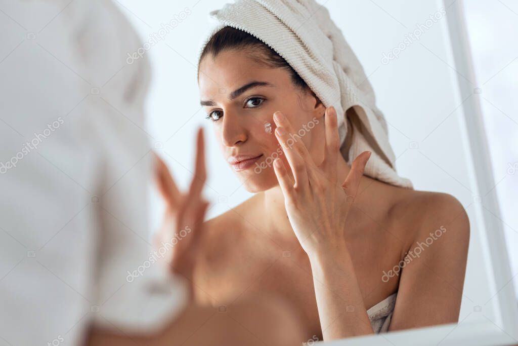 Portrait of beautiful young woman caring of her skin while putting on cream looking at mirror at home.