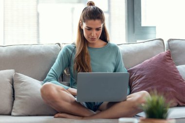Evde bir kanepede otururken dizüstü bilgisayarıyla çalışan kendine güvenen genç bir kadının fotoğrafı..