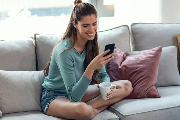 自宅のソファに座ってコーヒーを飲みながら携帯電話を使用してかなり若い笑顔の女性のショット — ストック写真