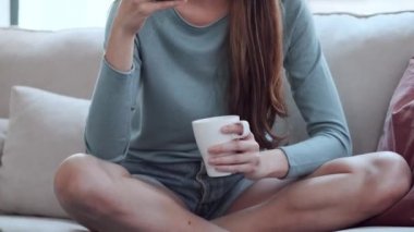 Evdeki bir kanepede oturup kahve içerken cep telefonuyla müzik dinleyen güzel ve genç bir kadının videosu..