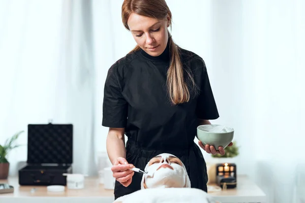 美容師のショットは スパセンターでストレッチャーに横たわっている間 女性にアルギン酸フェイスマスクを適用します — ストック写真