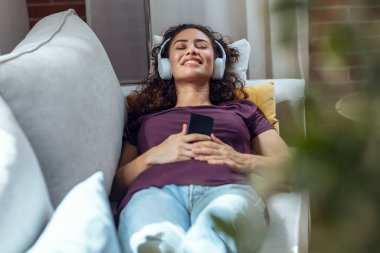 Evde kanepede uzanmış, akıllı telefonuyla müzik dinleyen rahat genç bir kadın..