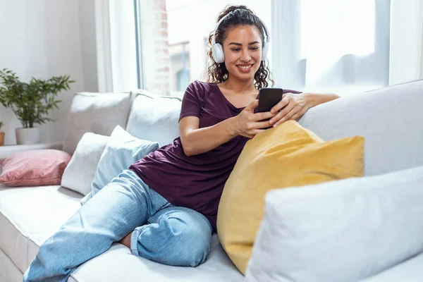 家でソファに座っている間にスマートフォンで音楽を聞いて笑顔の若い女性のショット — ストック写真