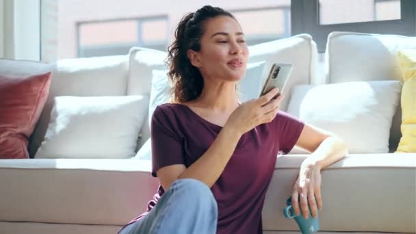 自宅の床に座ってラップトップで作業しながら 彼女の携帯電話を使用してかなり若い女性のビデオ — ストック動画