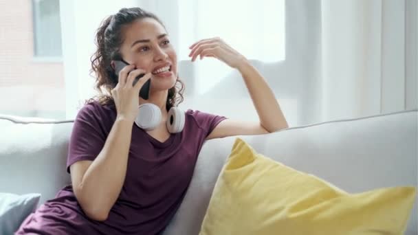家でソファに座っている間にスマートフォンで音楽を聴いている若い女性の笑顔のビデオ — ストック動画