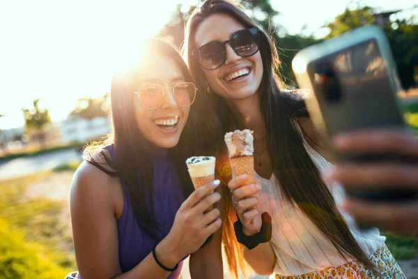 两名年轻貌美的女子在公园用手机拍照时吃冰淇淋 — 图库照片